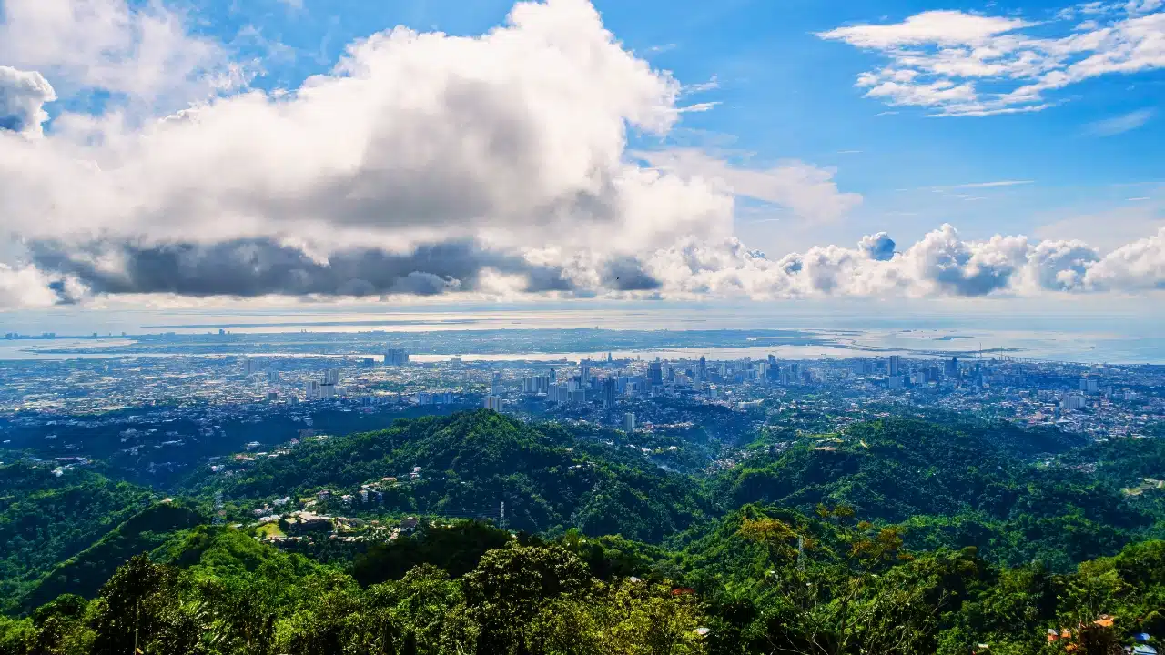 Cebu City skyline seen from mountain viewpoint with green hills, city buildings, and ocean coastline under dramatic clouds
